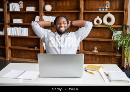 Smiling Afro-American businessman holding hands behind head sitting at office desk behind laptop. Happy black employee feeling no stress, relaxing, watching funny video after successful working Stock Photo