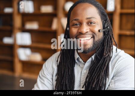 African american operator with long dreadlocks and headset, looking at camera, working in callcentre on support hotline in modern office with copy space, portrait of positive agent in conversation Stock Photo