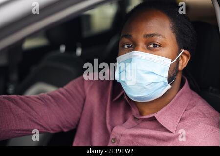 Black ethnic man driving, sitting in the car wearing surgical face mask looking out the window or at camera, driver with face mask protection against virus, close-up on mans face in respirator mask Stock Photo