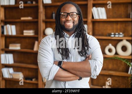 Portrait of a young strong african unshaved man with dreadlocks in glasses stands with arms crossed on his chest, looks at the camera, smiles, against the background of shelves with books, posing Stock Photo