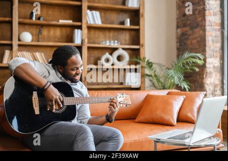 African man sitting on the leather sofa with guitar in a creative space or at home, playing a song for his girlfriend, teaching online, showing master class, recording song, learns song from a video Stock Photo
