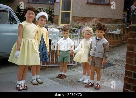 1960, historical, May and a group of small children standing in a suburban driveway in their sunday best clothes, two of the girls are wearing pretty yellow dresses, perhaps to attend a wedding or birthday party. The three youngest, two little boys and girl are holding each others hands, England, UK. Stock Photo