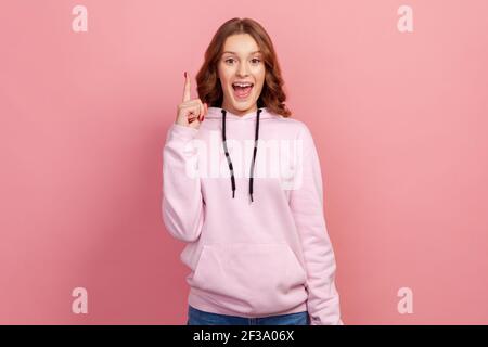 Portrait of excited inspired teen with brown hair pointing finger up looking at camera with toothy smile, happy with new great idea, find solution. In Stock Photo