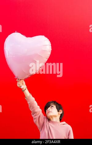 Boy holding a big pink heart shaped balloon against a red wall Stock Photo