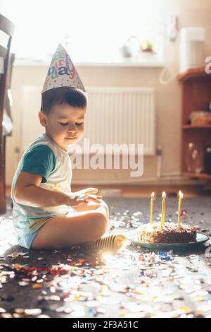 Close up photo of a small caucasian boy celebrating his birthday on the floor posing with a cake and confetti while wearing a holiday cap Stock Photo