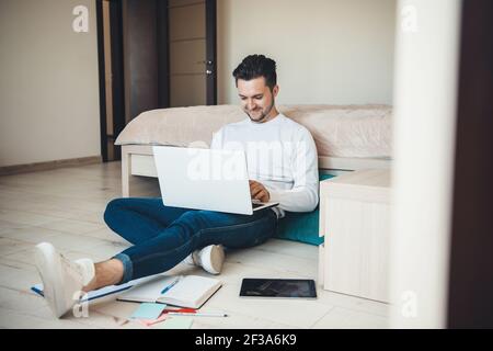 Smiling caucasian man is sitting on the floor working with a laptop remotely and some papers Stock Photo