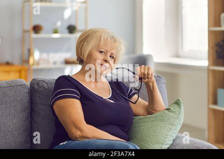 Wise mature woman holding glasses and looking at camera sitting on sofa at home Stock Photo
