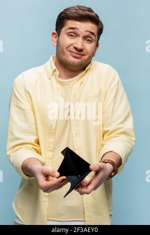 displeased man in yellow shirt holding black empty wallet isolated on blue Stock Photo