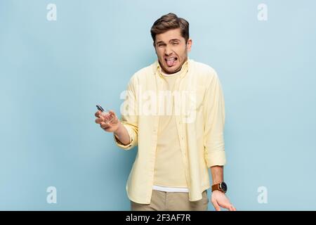disgusted young man in yellow shirt holding bottle with perfume and sticking out tongue on blue Stock Photo
