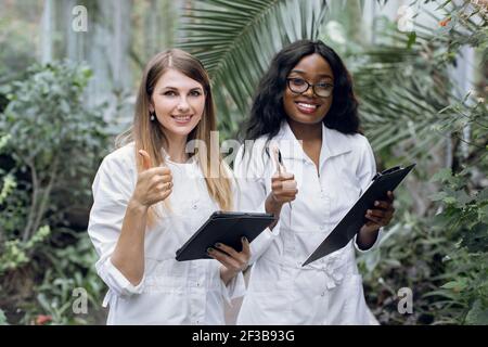 Team of young smiling two multiethnic women, professional scientists agronomists, posing to camera with smile and thumbs up, while standing in Stock Photo