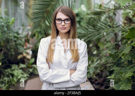 Portrait of young confident Caucasian woman, scientist, botanist, agronomist, wearing glasses and white lab coat, posing to camera with arms crossed Stock Photo