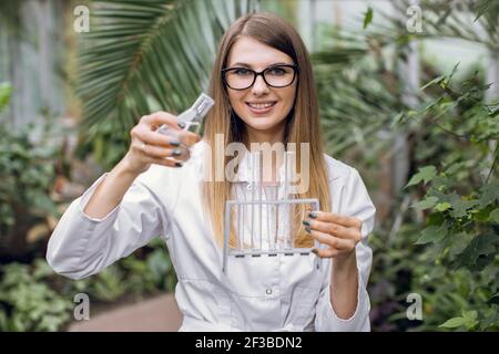 Young smiling likable woman engineer agronomist, working in a hydroponic garden, holding test tubes and flask, making scientific research, taking Stock Photo