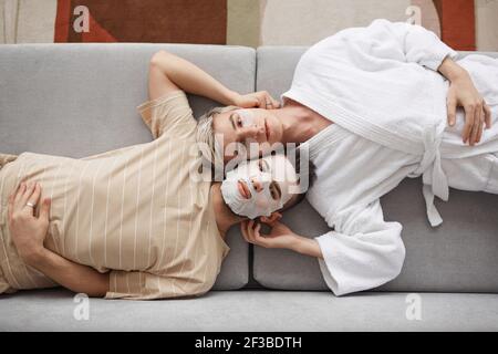Top view portrait of young gay couple looking at camera while lying on couch together and wearing face masks, beauty and skincare concept