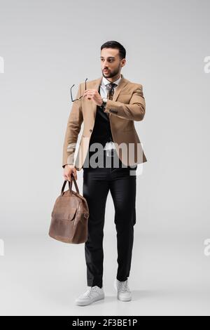 full length view of fashionable arabian man holding eyeglasses and leather bag on grey Stock Photo