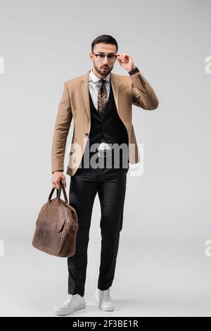 full length view of fashionable arabian man adjusting eyeglasses while posing with leather bag on grey Stock Photo