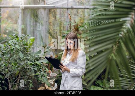 Professional female scientist biologist, pretty confident Caucasian woman in white coat, standing in greenhouse making notes while checking plants Stock Photo