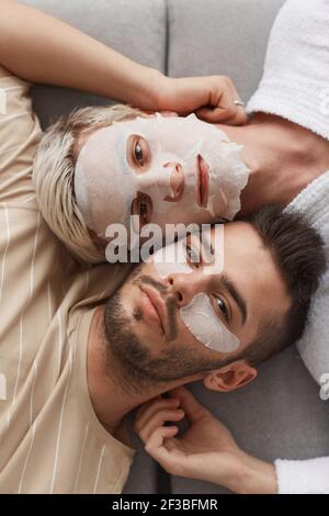 Top view closeup of young gay couple looking at camera while lying on couch together and wearing face masks, beauty and skincare concept