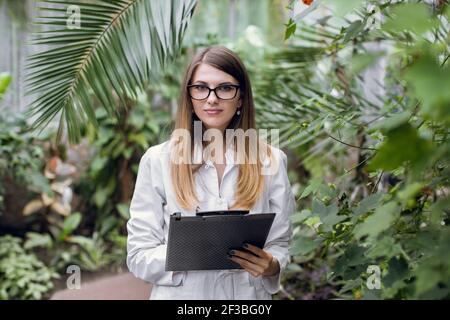 Greenhouse and tropic plants. Young attractive Caucasian woman scientist, posing to camera, while inspecting plants growing in greenhouse, holding a Stock Photo