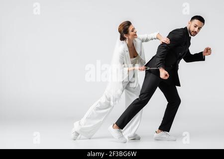Arabian man in formal wear and handcuffs running near bride on white background Stock Photo