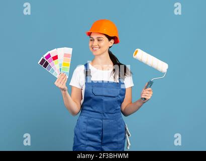 Smiling handywoman in coveralls and hardhat holding color palette and roller brush Stock Photo
