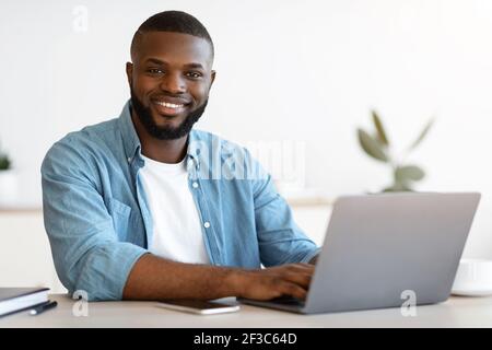 Portrait Of Happy Successful Black Freelancer Guy Posing At Desk With Laptop Stock Photo
