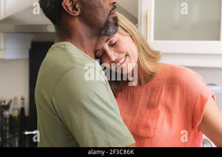 Diverse senior couple dancing in kitchen and embracing Stock Photo