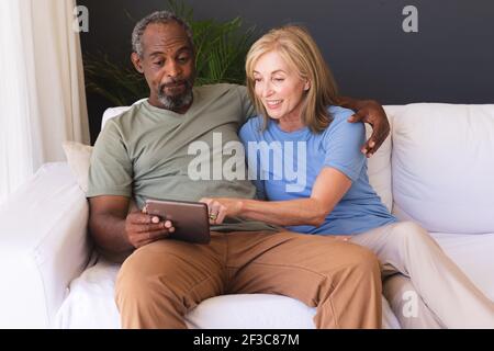 Diverse senior couple sitting on couch and using digital tablet Stock Photo
