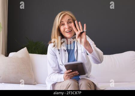 Caucasian senior female doctor sitting on sofa smiling giving video call consultation Stock Photo