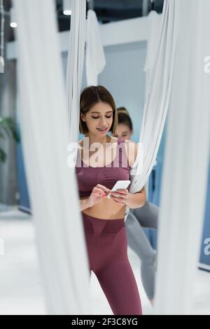 Young sportswoman using smartphone near fly yoga straps on blurred foreground Stock Photo
