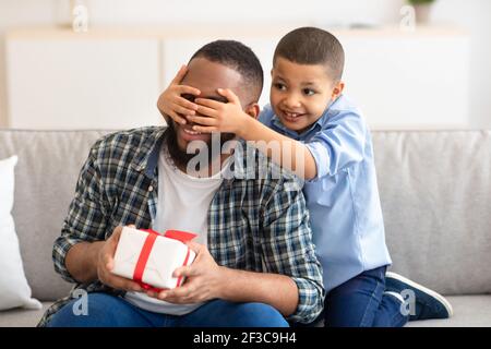 Black Son Congratulating Daddy Giving Gift Covering His Eyes Indoor Stock Photo