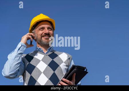 Smiling Business Person in Yellow Hardhat Talking on Mobile Phone. Stock Photo