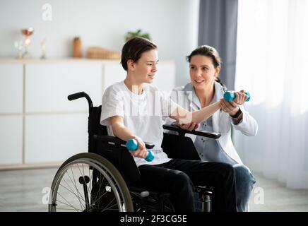 Physical rehabilitation concept. Young physiotherapist helping teenage boy in wheelchair to do exercises at home Stock Photo