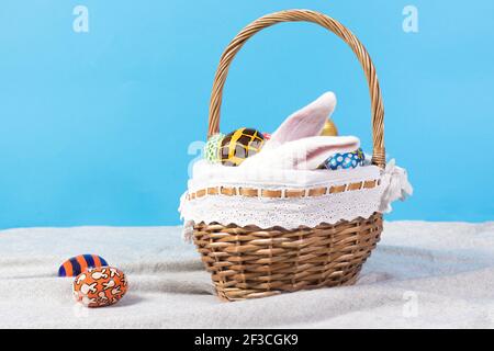 White long ears of a fluffy real rabbit in a basket on a blue background with Easter eggs. Easter holiday photo for postcard to copy space for banner. Stock Photo