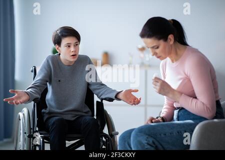 Family disagreement. Upset mother crying and her teenage son in wheelchair trying to justify himself at home Stock Photo
