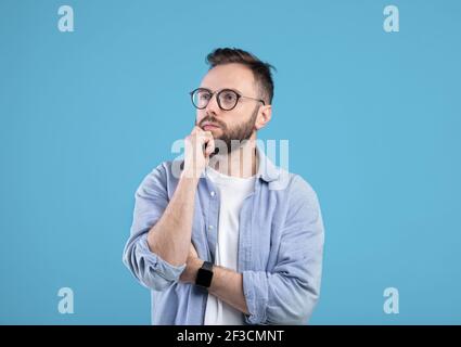 Portrait of dreamy Caucasian man in glasses touching his chin, thinking about something over blue studio background Stock Photo