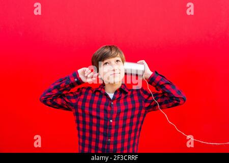 Boy using two metal cans and a thread as a telephone against a red wall Stock Photo