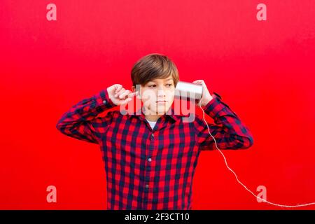 Boy using two metal cans and a thread as a telephone against a red wall Stock Photo