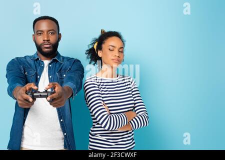 KYIV, UKRAINE - OCTOBER 23, 2020: offended african american woman standing with crossed arms near husband gaming on blue Stock Photo