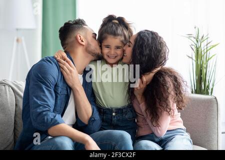 Family Love. Caring Parents Kissing Their Little Daughter, Bonding Together At Home Stock Photo