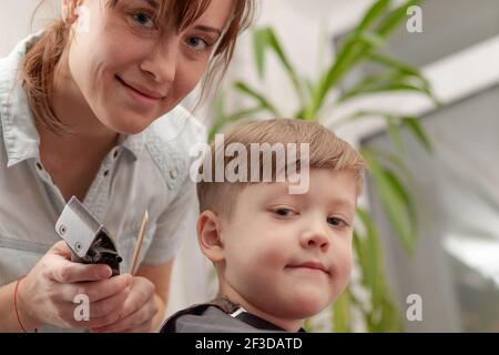 young mom hairdresser cuts her baby boy at home with hair clipper during quarantine. selective focus. portrait Stock Photo