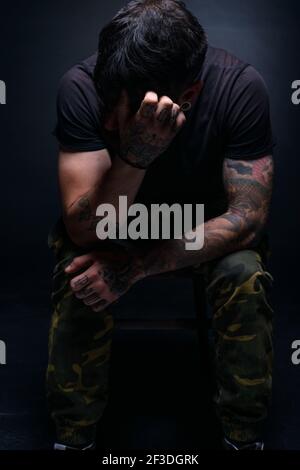 High contrast picture of a bearded male model sitting on a chair and covering his face with his tattooed hands Stock Photo