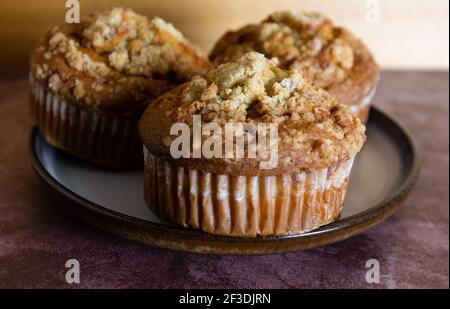 Side view of three cinnamon crumb muffins on a plate atop a maroon tabletop with a wall in the background. Stock Photo