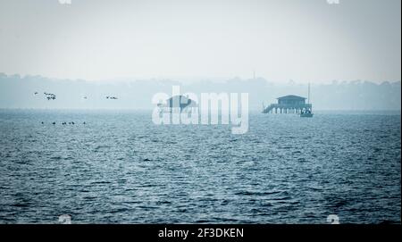 Hazy view of two wooden fisherman's houses on stilts. Winter seascape with rippled water. Coast disappearing in mist in background. Stock Photo