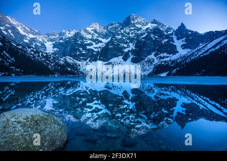 Reflecting of sharp rocky peaks dusted with snow in Eye of the sea (Morskie oko) lake in Poland. Winter in Tatra mountains. Stock Photo