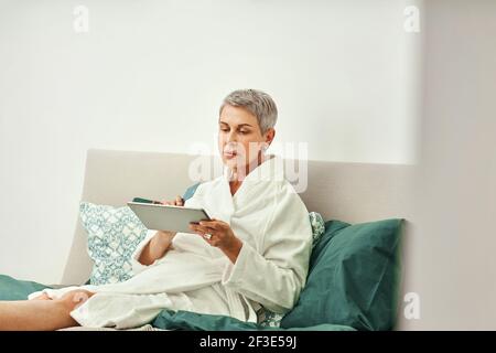 Mature woman relaxing at home. Senior female making online order while lying on a bed. Stock Photo