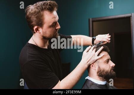 Professional barber doing haircut to bearded man customer using scissors and comb. Stock Photo