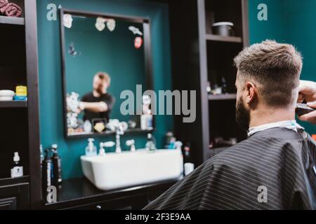 Bearded man getting haircut in barbershop, hairdresser cutting hair of male client with clipper. Stock Photo