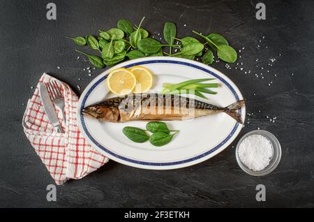 Whole smoked mackerel fish on white oval plate, gray black stone like table under, rock salt, lemon slices and green leaves salad near. View from abov Stock Photo
