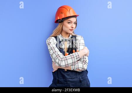 female engineer constructor hold tools for household in hands and looking at camera, in hardhat and blue uniform overalls. isolated blue studio backgr Stock Photo