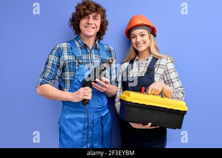 Two architects in uniform coveralls stand with tools instruments posing isolated on blue, young man hold drill in hands while his colleague woman hold Stock Photo
